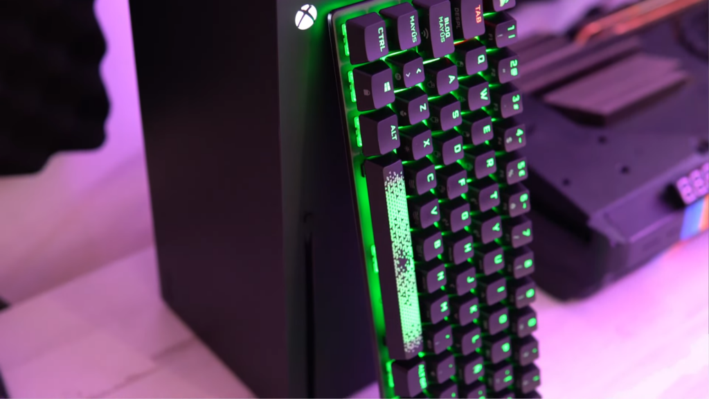 Se muestar el teclado apoyado sobre la Xbox series X y al lado la Playstation 5